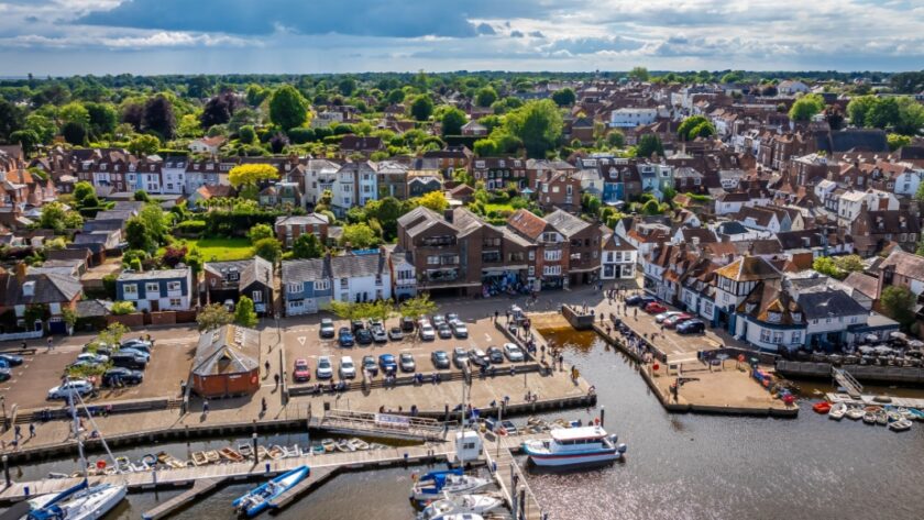 A scenic aerial view of Lymington harbour, showing moored boats and a waterfront lined with traditional buildings. Behind, rows of houses and green trees stretch into the distance under a partly cloudy sky