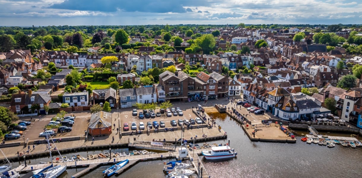 A scenic aerial view of Lymington harbour, showing moored boats and a waterfront lined with traditional buildings. Behind, rows of houses and green trees stretch into the distance under a partly cloudy sky