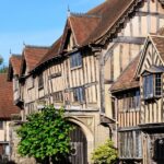 Historic timber-framed buildings in Warwick, England, including the Lord Leycester Hospital, with gabled roofs and leaded windows under a bright blue sky. The medieval architecture is well-preserved and richly detailed, showcasing the town’s centuries-old character.