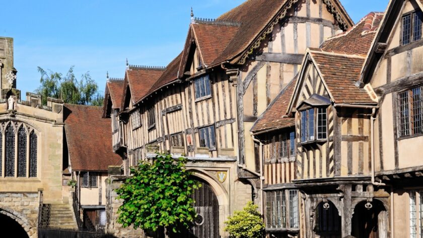 Historic timber-framed buildings in Warwick, England, including the Lord Leycester Hospital, with gabled roofs and leaded windows under a bright blue sky. The medieval architecture is well-preserved and richly detailed, showcasing the town’s centuries-old character.