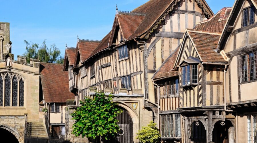 Historic timber-framed buildings in Warwick, England, including the Lord Leycester Hospital, with gabled roofs and leaded windows under a bright blue sky. The medieval architecture is well-preserved and richly detailed, showcasing the town’s centuries-old character.