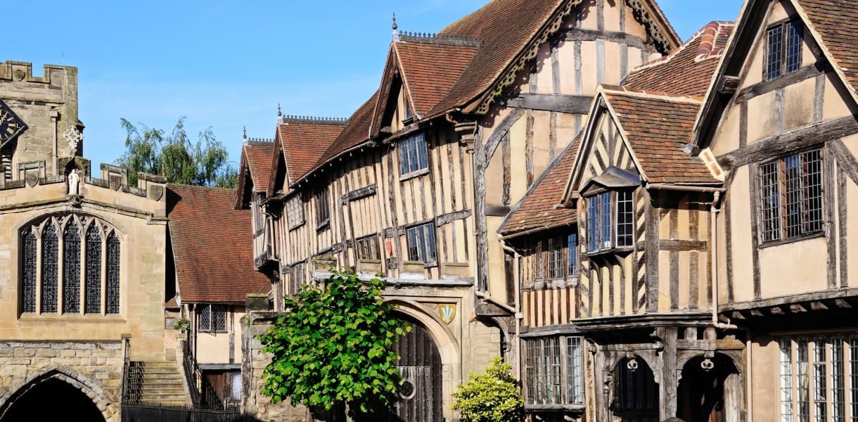 Historic timber-framed buildings in Warwick, England, including the Lord Leycester Hospital, with gabled roofs and leaded windows under a bright blue sky. The medieval architecture is well-preserved and richly detailed, showcasing the town’s centuries-old character.