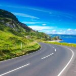 Sweeping coastal road curving past lush green cliffs beside a vivid blue sea under a bright summer sky in Northern Ireland.