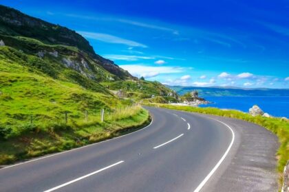 Sweeping coastal road curving past lush green cliffs beside a vivid blue sea under a bright summer sky in Northern Ireland.