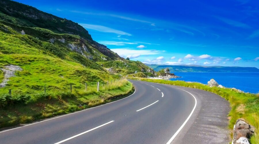 Sweeping coastal road curving past lush green cliffs beside a vivid blue sea under a bright summer sky in Northern Ireland.