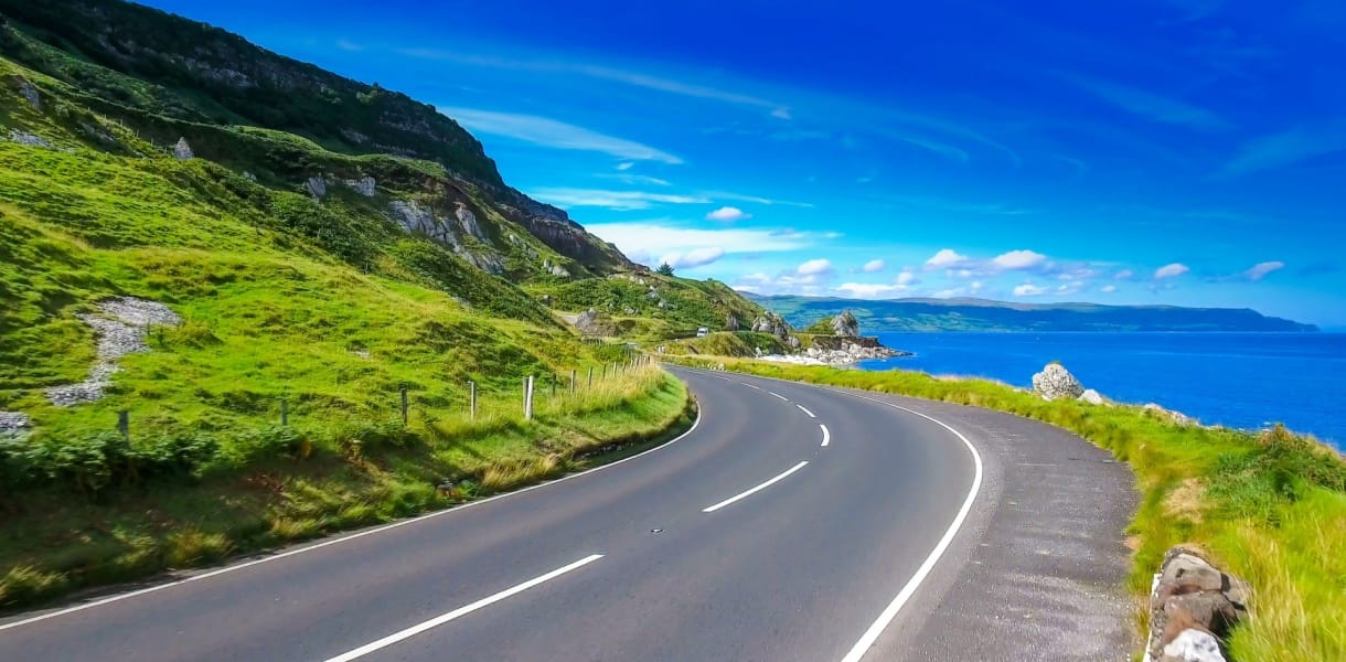 Sweeping coastal road curving past lush green cliffs beside a vivid blue sea under a bright summer sky in Northern Ireland.