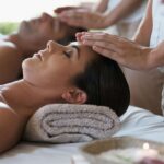 A woman and man lying side by side on massage beds receiving head massages at a luxury hotel spa, surrounded by soft towels and calming decor.