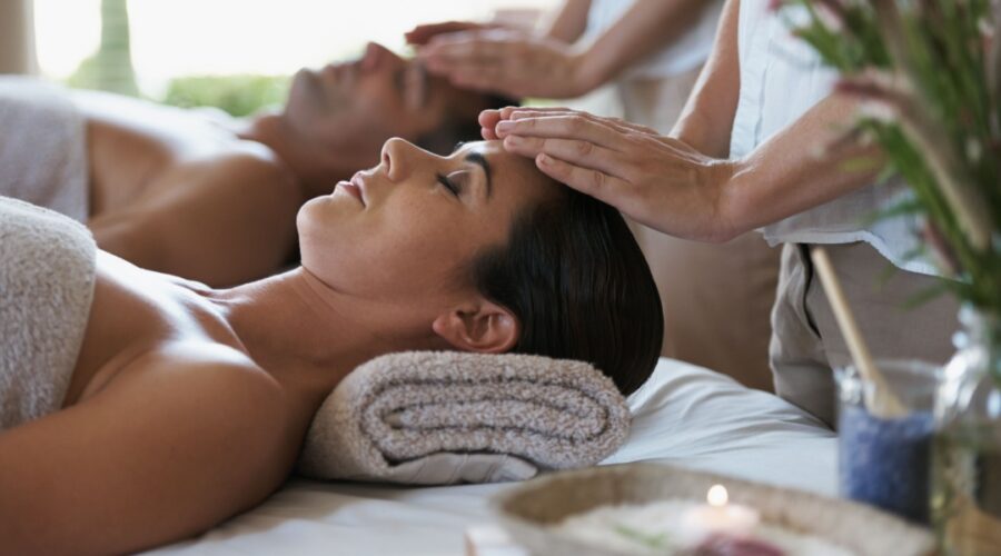 A woman and man lying side by side on massage beds receiving head massages at a luxury hotel spa, surrounded by soft towels and calming decor.