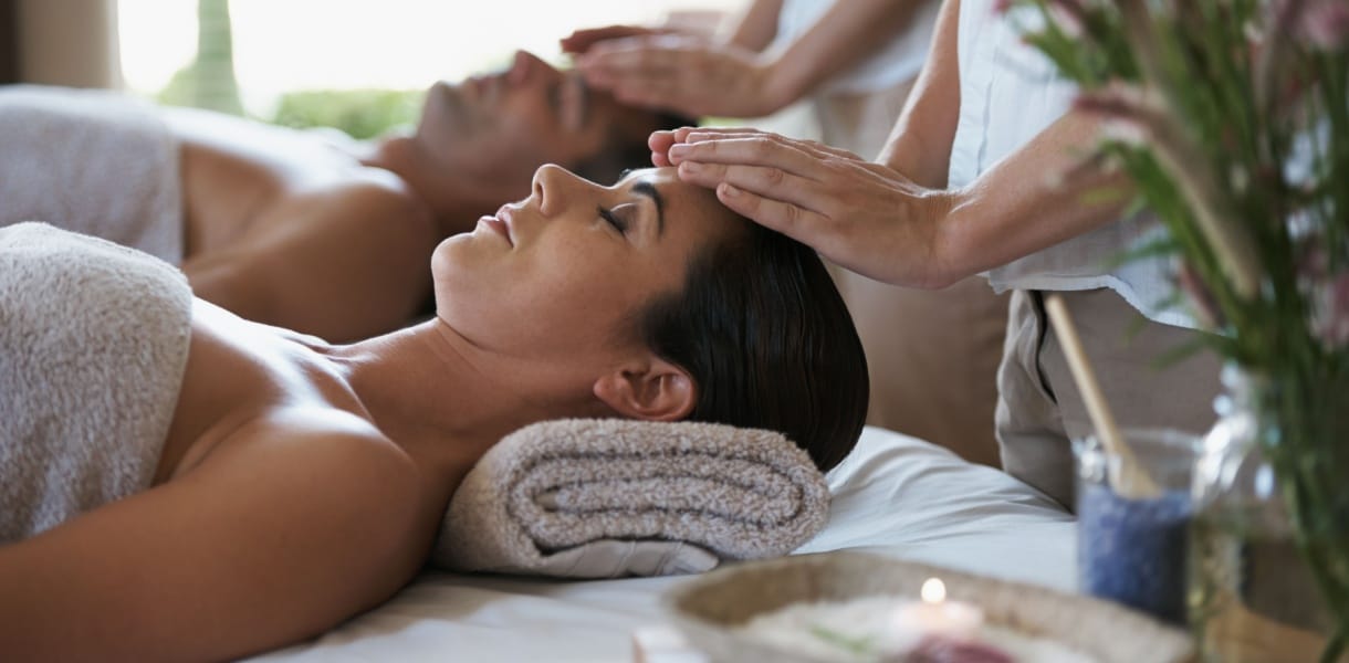 A woman and man lying side by side on massage beds receiving head massages at a luxury hotel spa, surrounded by soft towels and calming decor.
