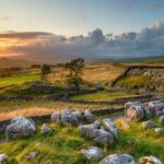 Sunset over the rolling hills and dry-stone walls of the Yorkshire Dales, with rocky foreground, winding dry stone walls, and a golden sky casting warm light across the landscape