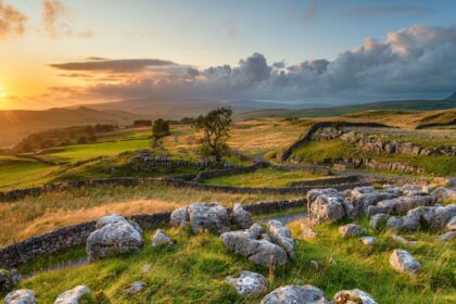 Sunset over the rolling hills and dry-stone walls of the Yorkshire Dales, with rocky foreground, winding dry stone walls, and a golden sky casting warm light across the landscape