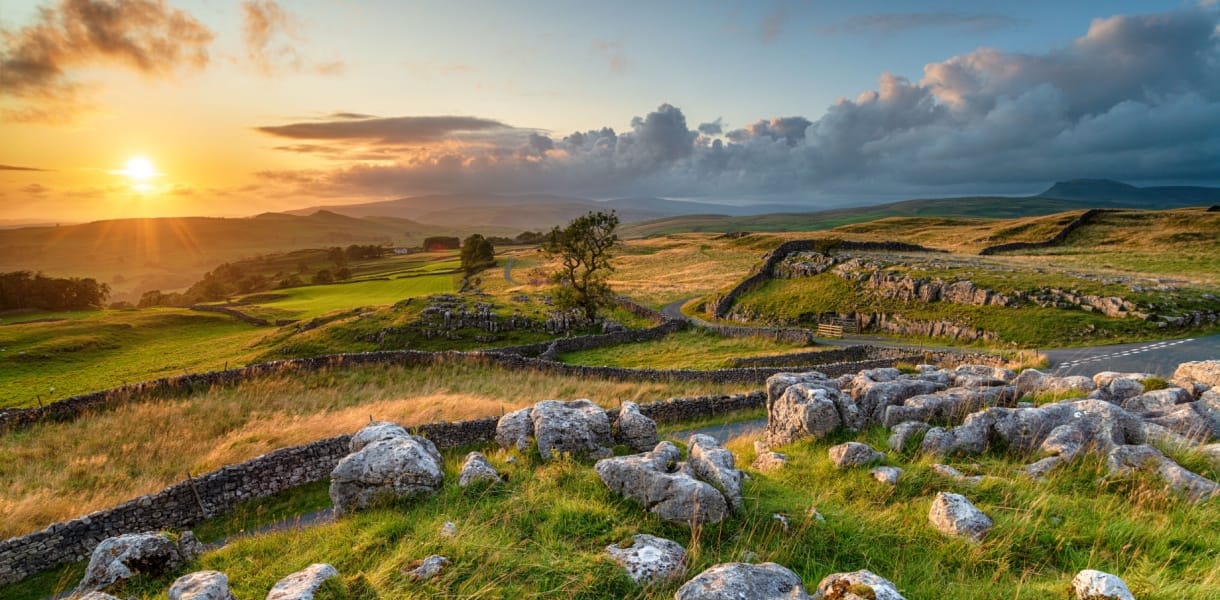 Sunset over the rolling hills and dry-stone walls of the Yorkshire Dales, with rocky foreground, winding dry stone walls, and a golden sky casting warm light across the landscape