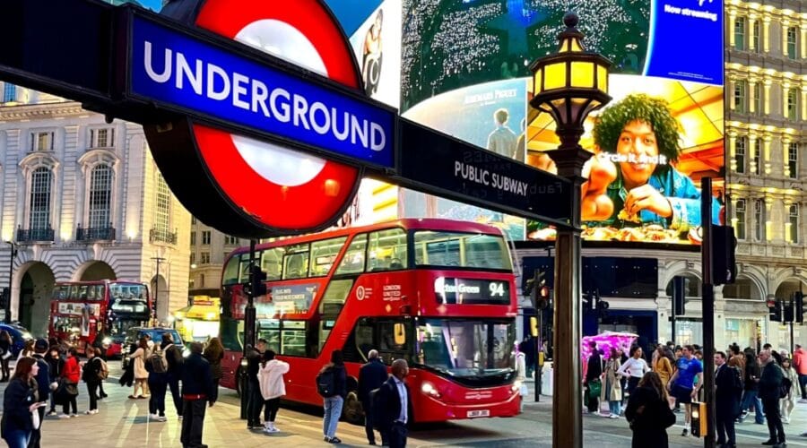 London, Piccadilly Circus at dusk with crowds, red double-decker buses, and illuminated billboards, including a bright Underground sign in the foreground.