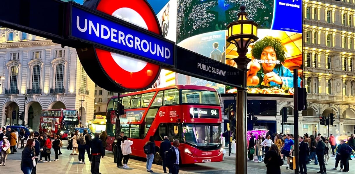 London, Piccadilly Circus at dusk with crowds, red double-decker buses, and illuminated billboards, including a bright Underground sign in the foreground.