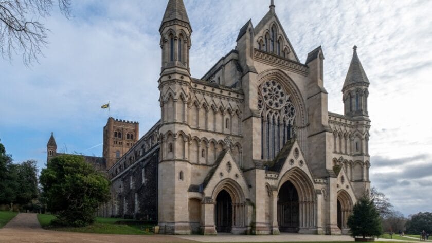 St Albans Cathedral in Hertfordshire, England, viewed from the west front. The grand Norman and Gothic façade features tall towers, pointed spires, and an ornate rose window above three arched doorways. To the left, the central tower rises with a flag flying, surrounded by trees and greenery under a partly cloudy sky.