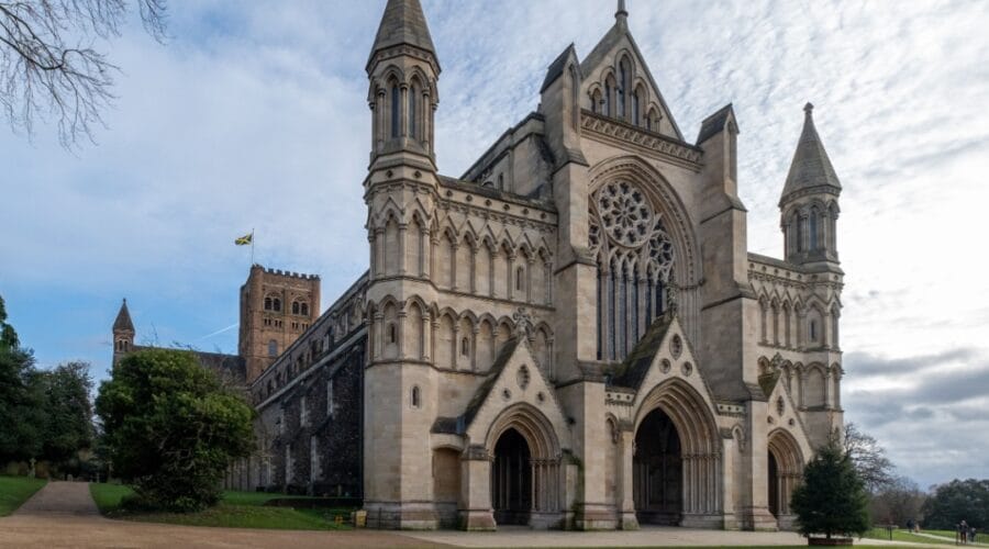 St Albans Cathedral in Hertfordshire, England, viewed from the west front. The grand Norman and Gothic façade features tall towers, pointed spires, and an ornate rose window above three arched doorways. To the left, the central tower rises with a flag flying, surrounded by trees and greenery under a partly cloudy sky.