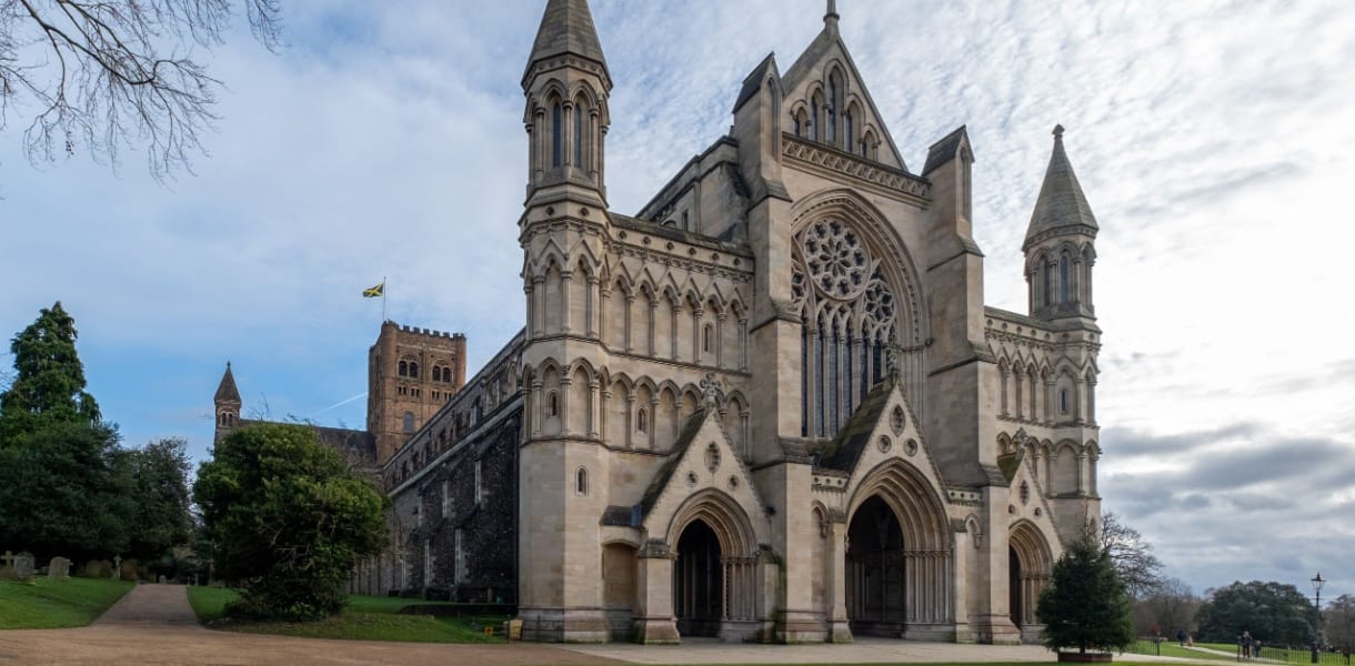 St Albans Cathedral in Hertfordshire, England, viewed from the west front. The grand Norman and Gothic façade features tall towers, pointed spires, and an ornate rose window above three arched doorways. To the left, the central tower rises with a flag flying, surrounded by trees and greenery under a partly cloudy sky.