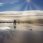 Two people walk along the reflective wet sands of Beadnell Bay under a dramatic sky, with sunbeams breaking through the clouds and casting a golden glow across the beach and distant dunes