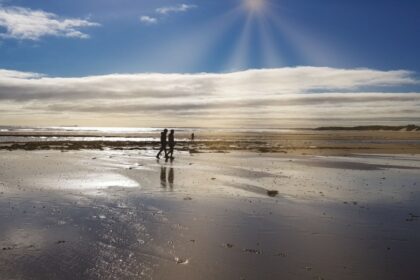 Two people walk along the reflective wet sands of Beadnell Bay under a dramatic sky, with sunbeams breaking through the clouds and casting a golden glow across the beach and distant dunes