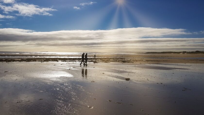 Two people walk along the reflective wet sands of Beadnell Bay under a dramatic sky, with sunbeams breaking through the clouds and casting a golden glow across the beach and distant dunes