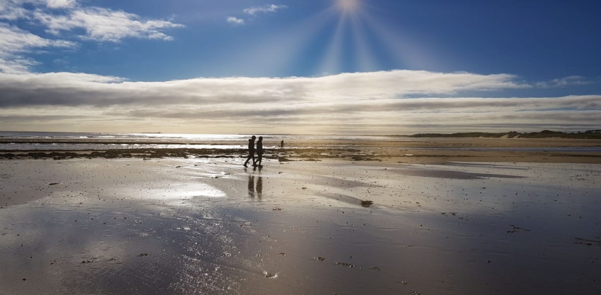 Two people walk along the reflective wet sands of Beadnell Bay under a dramatic sky, with sunbeams breaking through the clouds and casting a golden glow across the beach and distant dunes
