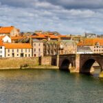 View of Berwick-upon-Tweed with the historic Old Bridge crossing the River Tweed, surrounded by red-roofed buildings and a church clock tower under a dramatic cloudy sky