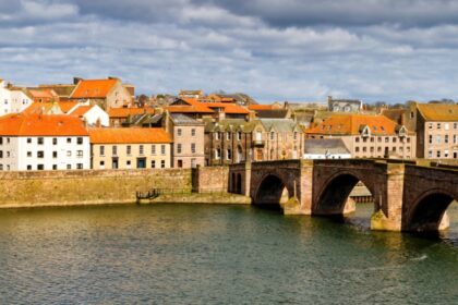 View of Berwick-upon-Tweed with the historic Old Bridge crossing the River Tweed, surrounded by red-roofed buildings and a church clock tower under a dramatic cloudy sky