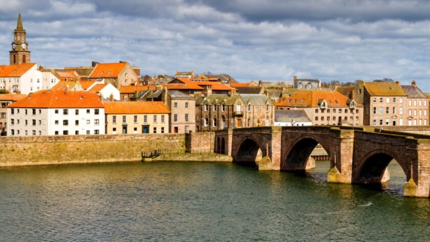 View of Berwick-upon-Tweed with the historic Old Bridge crossing the River Tweed, surrounded by red-roofed buildings and a church clock tower under a dramatic cloudy sky