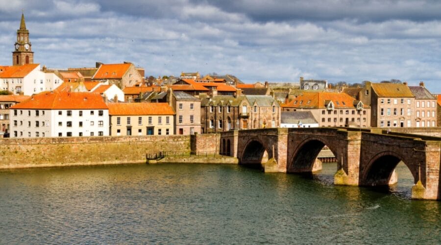 View of Berwick-upon-Tweed with the historic Old Bridge crossing the River Tweed, surrounded by red-roofed buildings and a church clock tower under a dramatic cloudy sky