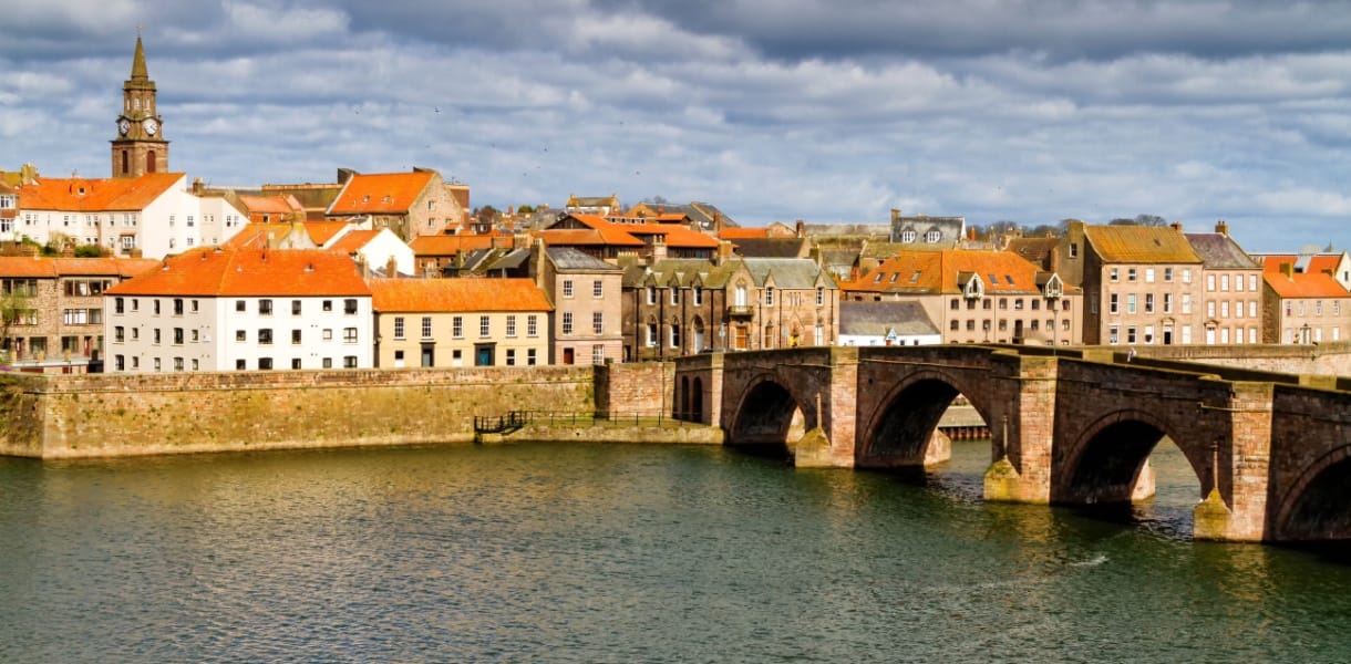 View of Berwick-upon-Tweed with the historic Old Bridge crossing the River Tweed, surrounded by red-roofed buildings and a church clock tower under a dramatic cloudy sky