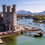 Aerial view of Caernarfon Castle in North Wales, with its towering stone walls overlooking the Menai Strait, surrounded by boats on the water and distant mountains under a clear blue sky.
