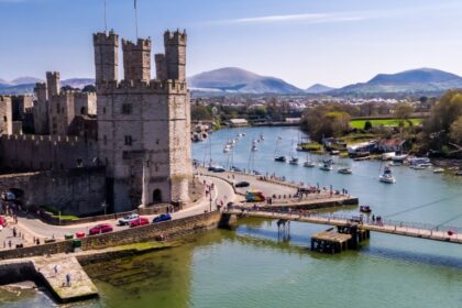 Aerial view of Caernarfon Castle in North Wales, with its towering stone walls overlooking the Menai Strait, surrounded by boats on the water and distant mountains under a clear blue sky.
