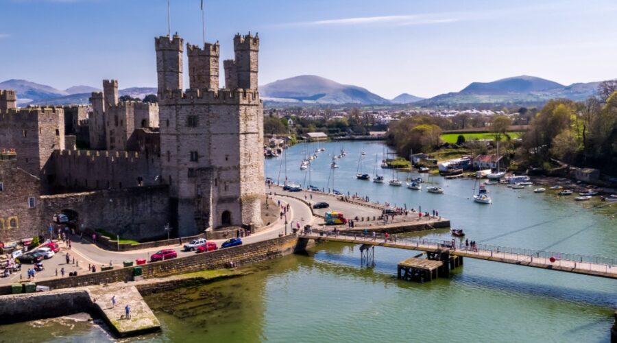 Aerial view of Caernarfon Castle in North Wales, with its towering stone walls overlooking the Menai Strait, surrounded by boats on the water and distant mountains under a clear blue sky.