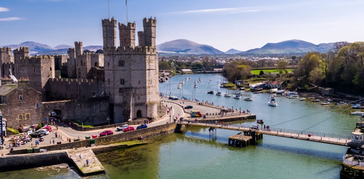 Aerial view of Caernarfon Castle in North Wales, with its towering stone walls overlooking the Menai Strait, surrounded by boats on the water and distant mountains under a clear blue sky.