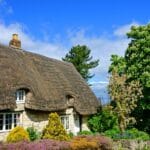 Traditional Cotswold stone cottage in Gloucestershire with a thatched roof, white-framed windows, and a colourful front garden, set beneath a bright blue sky with fluffy clouds and surrounded by lush green trees.