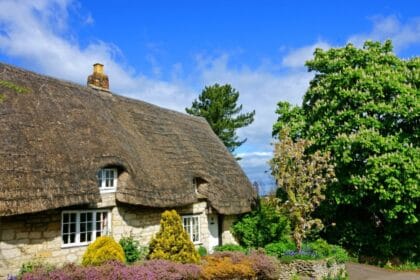 Traditional Cotswold stone cottage in Gloucestershire with a thatched roof, white-framed windows, and a colourful front garden, set beneath a bright blue sky with fluffy clouds and surrounded by lush green trees.