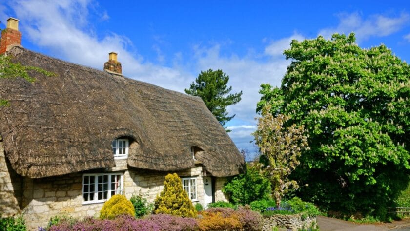 Traditional Cotswold stone cottage in Gloucestershire with a thatched roof, white-framed windows, and a colourful front garden, set beneath a bright blue sky with fluffy clouds and surrounded by lush green trees.