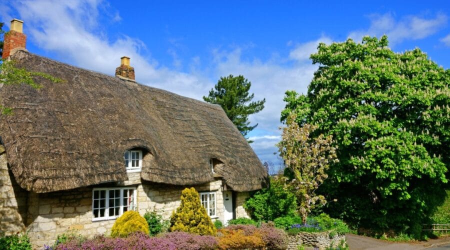 Traditional Cotswold stone cottage in Gloucestershire with a thatched roof, white-framed windows, and a colourful front garden, set beneath a bright blue sky with fluffy clouds and surrounded by lush green trees.