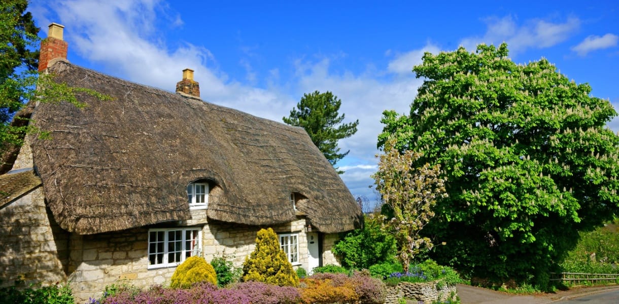 Traditional Cotswold stone cottage in Gloucestershire with a thatched roof, white-framed windows, and a colourful front garden, set beneath a bright blue sky with fluffy clouds and surrounded by lush green trees.