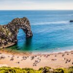 A sunny day at Durdle Door on the Dorset coast, with people relaxing on the sandy beach and swimming in the calm, turquoise sea beneath the famous limestone arch.