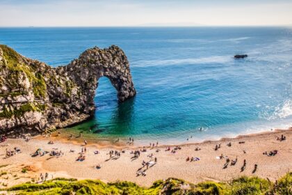 A sunny day at Durdle Door on the Dorset coast, with people relaxing on the sandy beach and swimming in the calm, turquoise sea beneath the famous limestone arch.