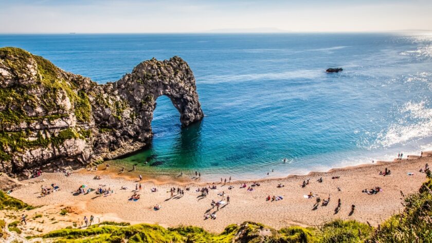A sunny day at Durdle Door on the Dorset coast, with people relaxing on the sandy beach and swimming in the calm, turquoise sea beneath the famous limestone arch.