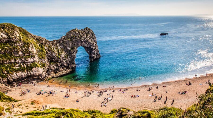 A sunny day at Durdle Door on the Dorset coast, with people relaxing on the sandy beach and swimming in the calm, turquoise sea beneath the famous limestone arch.