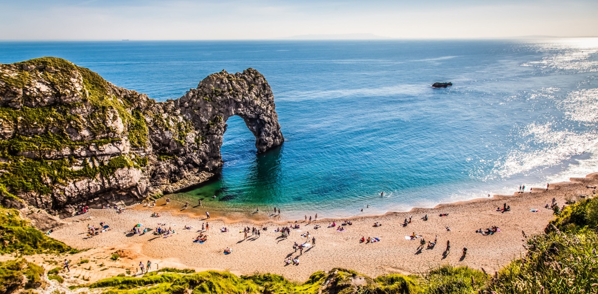 A sunny day at Durdle Door on the Dorset coast, with people relaxing on the sandy beach and swimming in the calm, turquoise sea beneath the famous limestone arch.