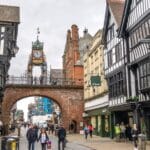 People walking along Eastgate Street beneath the historic Eastgate Clock in Chester, Cheshire, with black-and-white timber-framed buildings and shops lining the cobbled street.