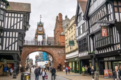 People walking along Eastgate Street beneath the historic Eastgate Clock in Chester, Cheshire, with black-and-white timber-framed buildings and shops lining the cobbled street.