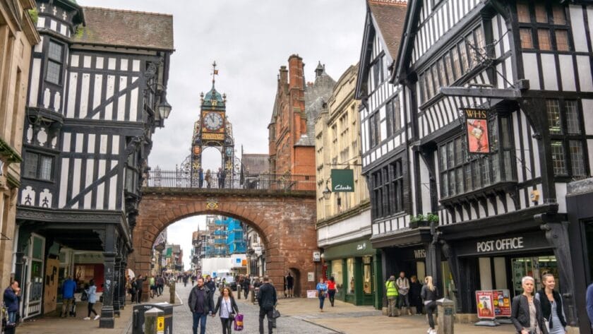 People walking along Eastgate Street beneath the historic Eastgate Clock in Chester, Cheshire, with black-and-white timber-framed buildings and shops lining the cobbled street.