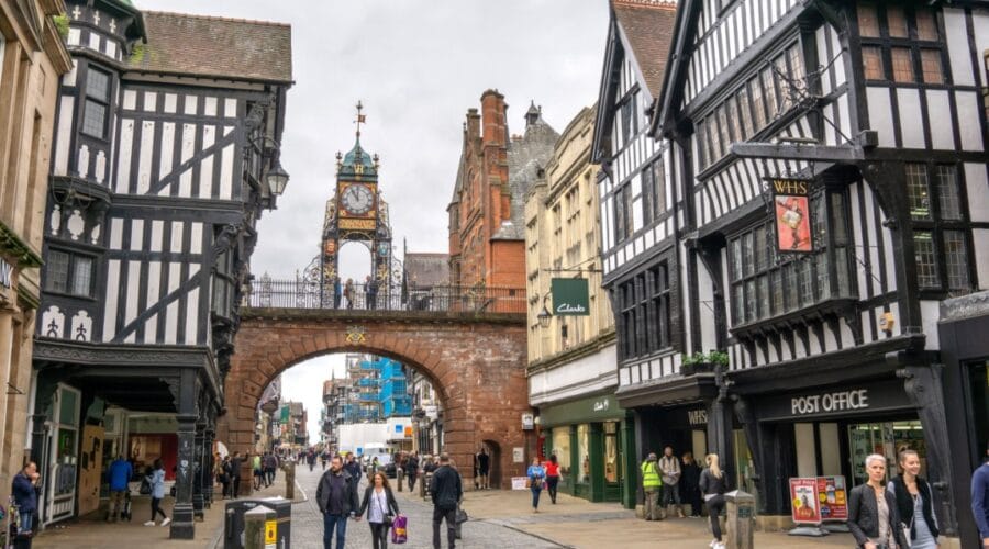 People walking along Eastgate Street beneath the historic Eastgate Clock in Chester, Cheshire, with black-and-white timber-framed buildings and shops lining the cobbled street.