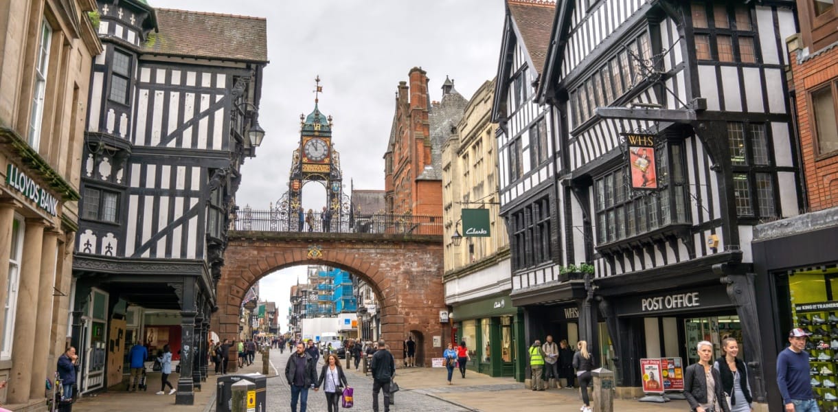 People walking along Eastgate Street beneath the historic Eastgate Clock in Chester, Cheshire, with black-and-white timber-framed buildings and shops lining the cobbled street.