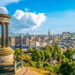 View of Edinburgh from Calton Hill on a sunny day, showing the Dugald Stewart Monument in the foreground and a panoramic skyline featuring Edinburgh Castle, the Balmoral Hotel clock tower, and the spires of the Old Town, with the Pentland Hills in the background.